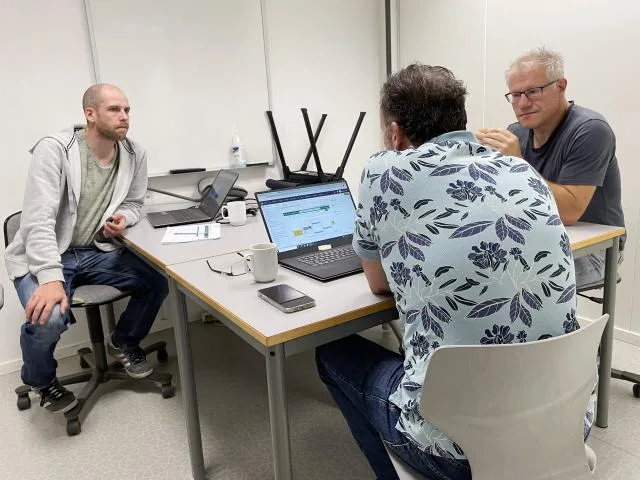 Tre menn sitter i diskusjon i klasserom foran en pc-skjerm.